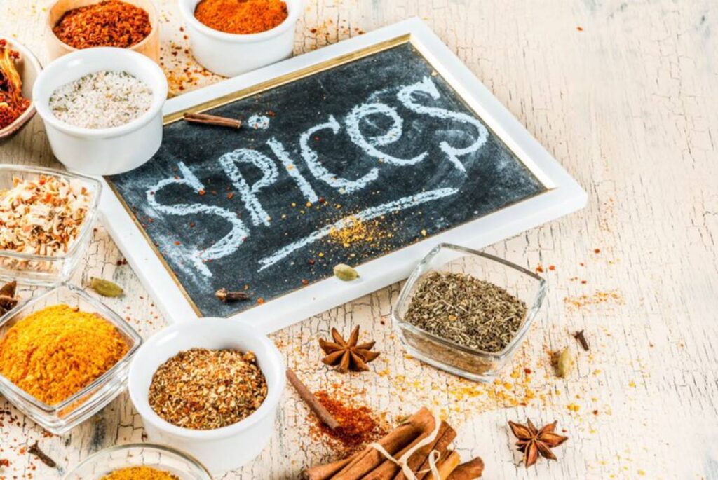 A chalkboard labeled "spices" surrounded by various spice containers, including herbs, powders, and whole spices on a rustic table.