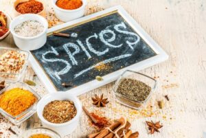 A chalkboard labeled "spices" surrounded by various spice containers, including herbs, powders, and whole spices on a rustic table.
