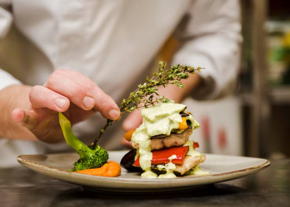 A chef delicately garnishes a vibrant, stacked vegetable dish with a sprig of thyme and an artistic drizzle of sauce.