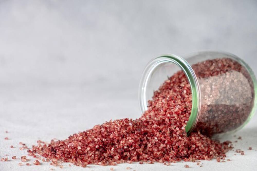 A glass jar tipped over, spilling coarse Red Alaea Salt onto a light surface, creating a textured mound and capturing a serene, minimalist vibe.