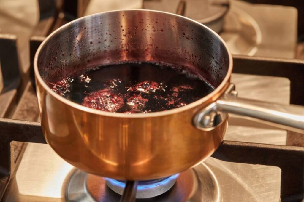 A copper saucepan simmering with dark purple liquid on a gas stove, bubbles forming and steam rising from the surface.