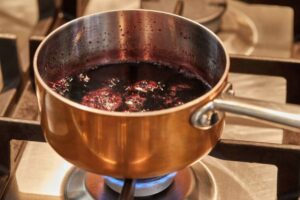 A copper saucepan simmering with dark purple liquid on a gas stove, bubbles forming and steam rising from the surface.