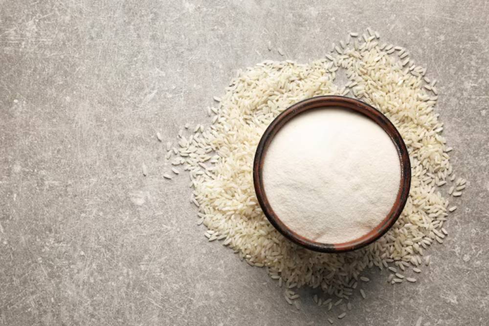 A wooden bowl filled with white flour surrounded by scattered uncooked white rice on a textured gray surface.