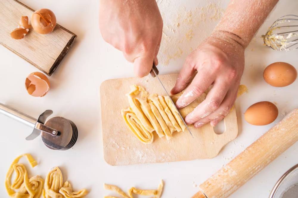 A person cuts freshly made pasta on a wooden board, surrounded by flour, eggs, and kitchen tools.