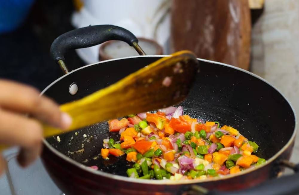 A hand stirs colorful diced vegetables in a frying pan with a wooden spatula, set against a blurred kitchen background.