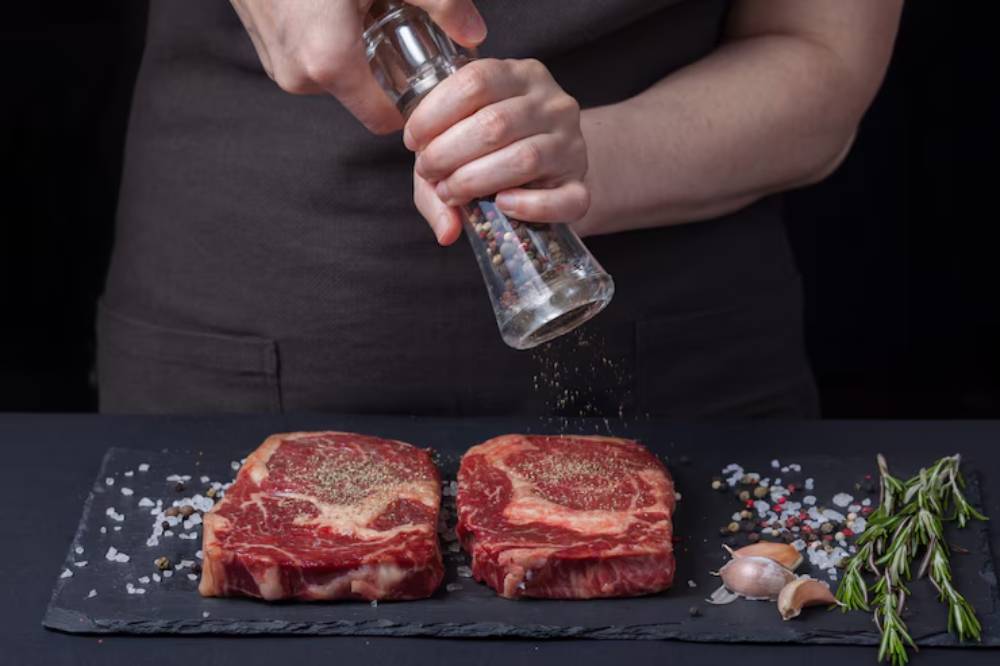 A hand seasoning two raw ribeye steaks with mixed peppercorns, alongside salt, garlic, and rosemary on a slate board.