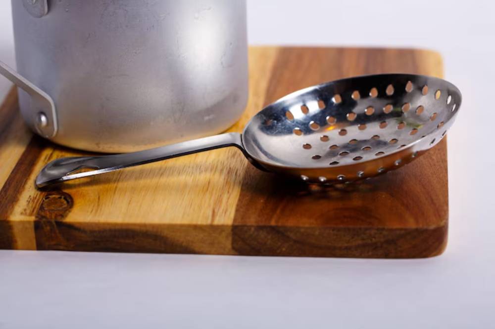 A silver slotted spoon rests on a wooden cutting board beside a metal pot, ready for cooking tasks.