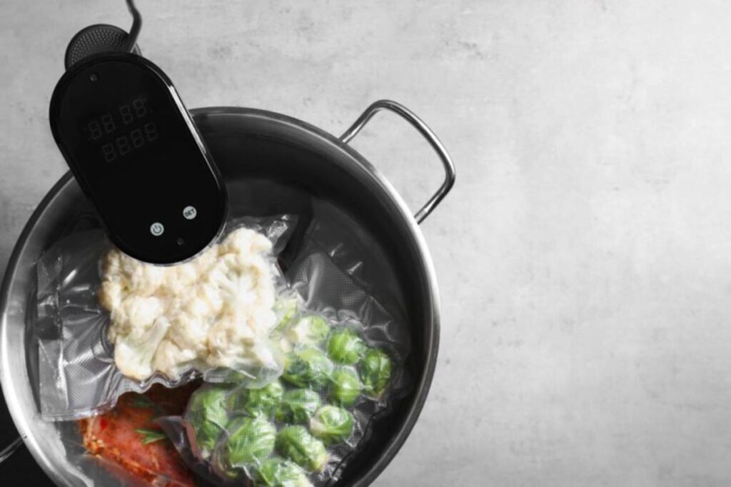 A sous vide device attached to a pot, with vacuum-sealed bags of cauliflower, leafy greens, and a red sauce submerged in water.
