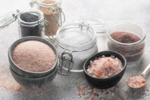 An assortment of salts in jars and bowls, including pink Himalayan, gray sea salt, and black salt, arranged artistically on a gray surface.
