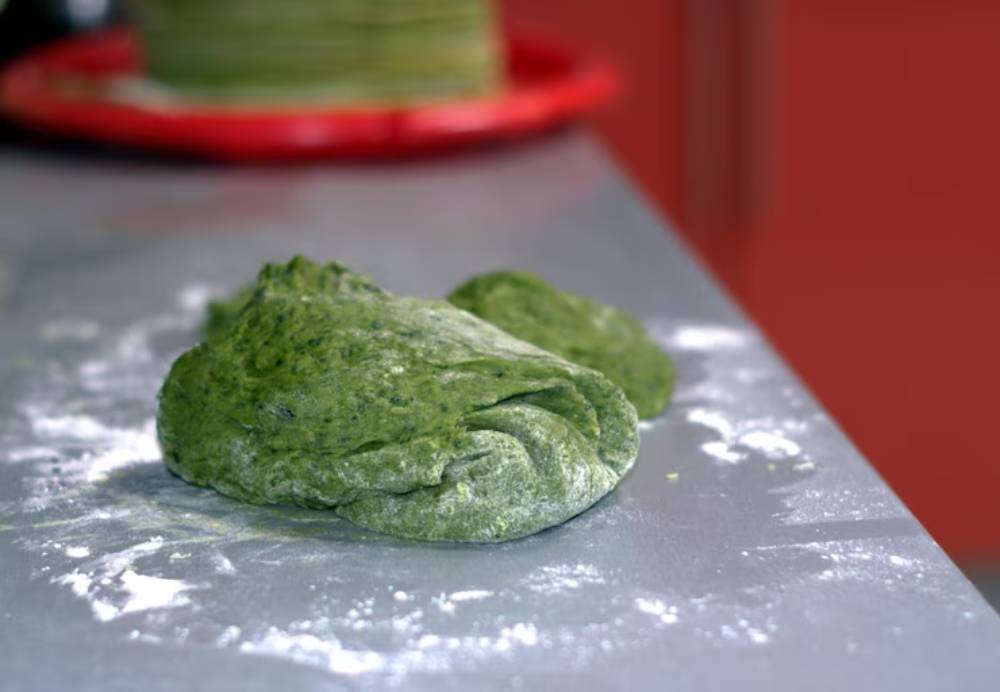 A ball of green spinach dough rests on a floured countertop, with a blurred background of stacked pasta and a red plate.
