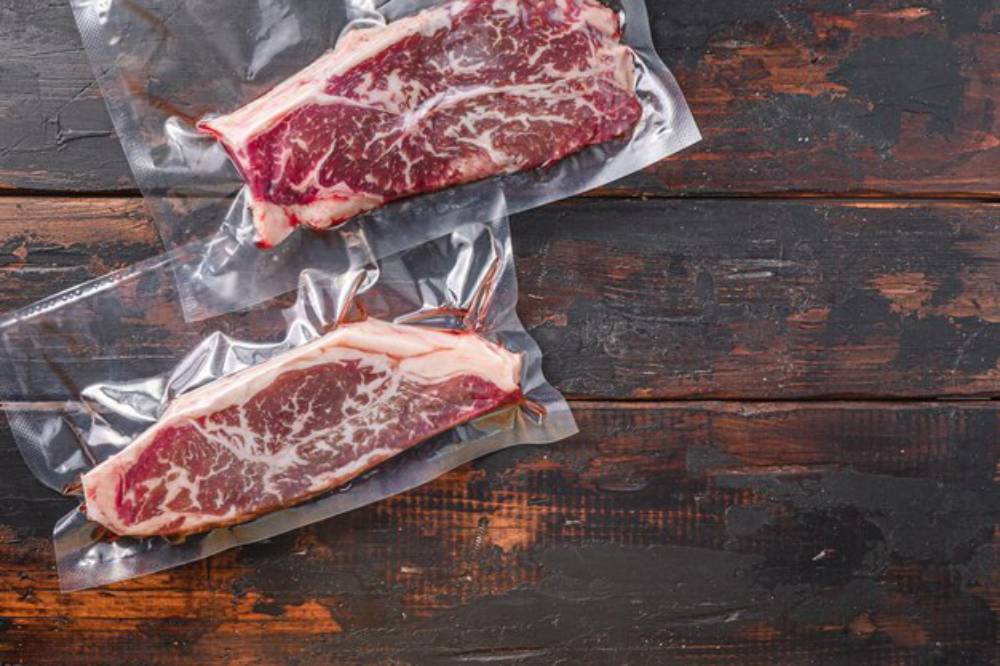 Two vacuum-sealed ribeye steaks are laid on a rustic wooden surface, showcasing marbled meat textures.