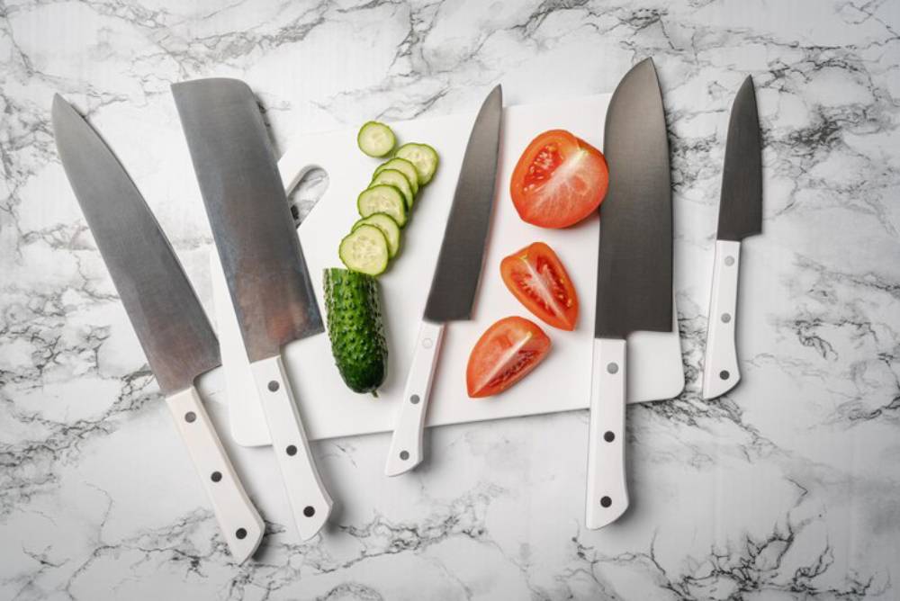 A collection of five knives rests on a white cutting board, surrounded by sliced cucumbers and tomatoes on a marble surface.