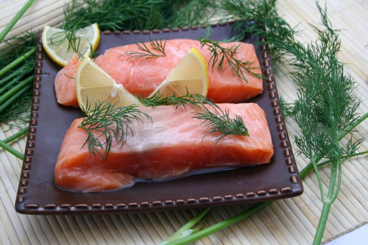 Two fresh salmon fillets garnished with dill and lemon wedges, displayed on a brown plate, surrounded by green herbs.