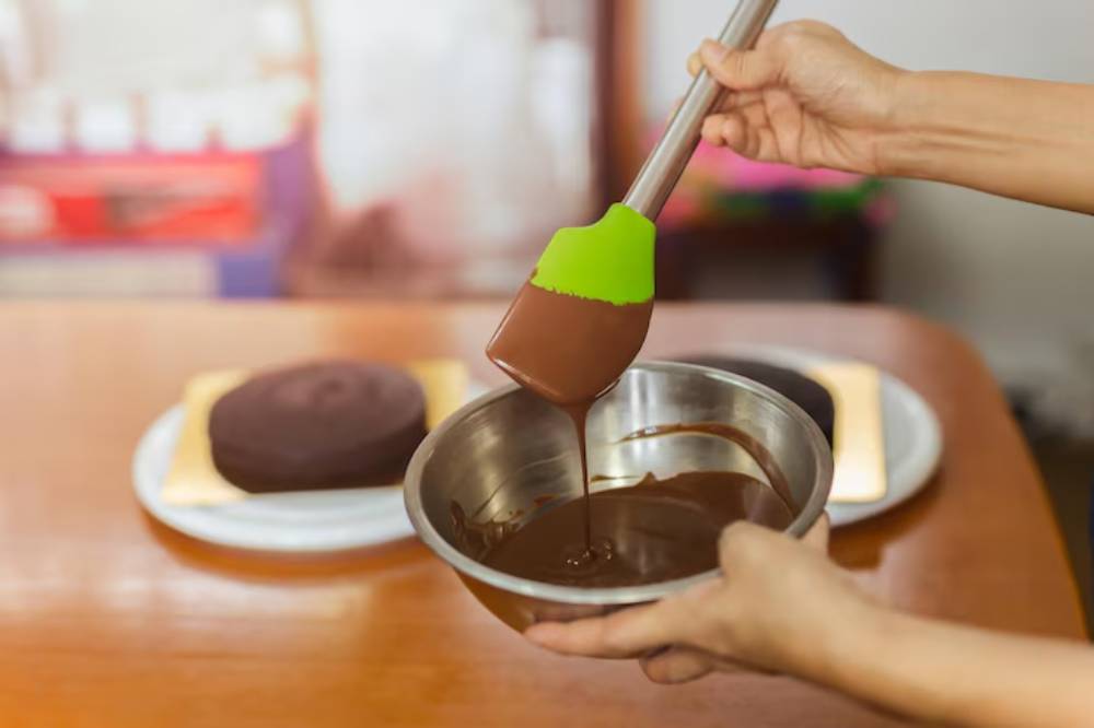 A person's hand lifts a spoonful of smooth chocolate from a bowl, with two round cakes on a plate in the background.