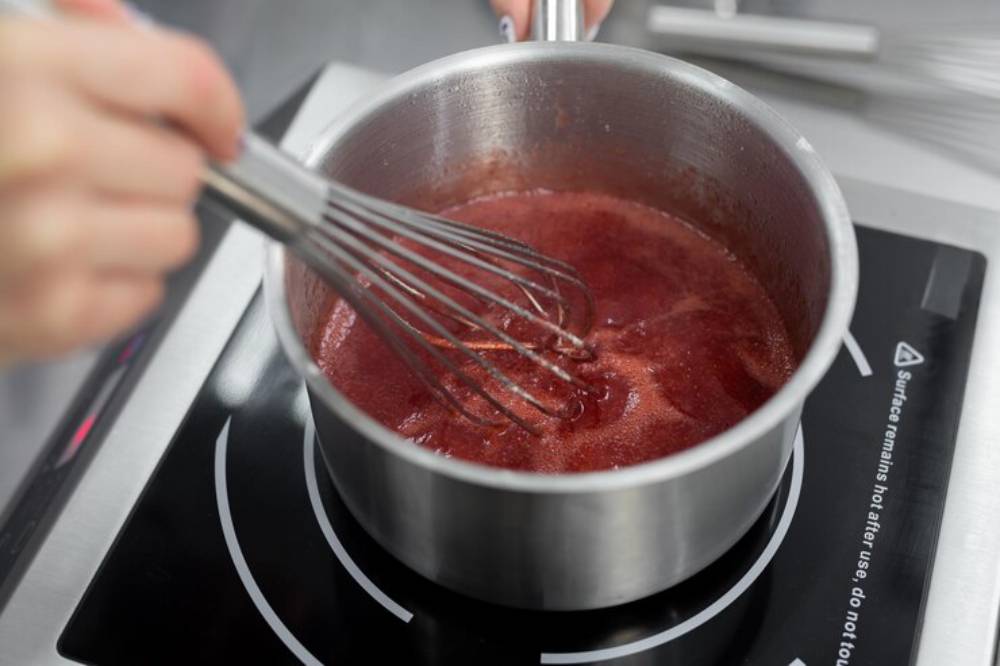 A hand with a whisk stirs a simmering red sauce in a stainless steel pot on an induction cooktop.