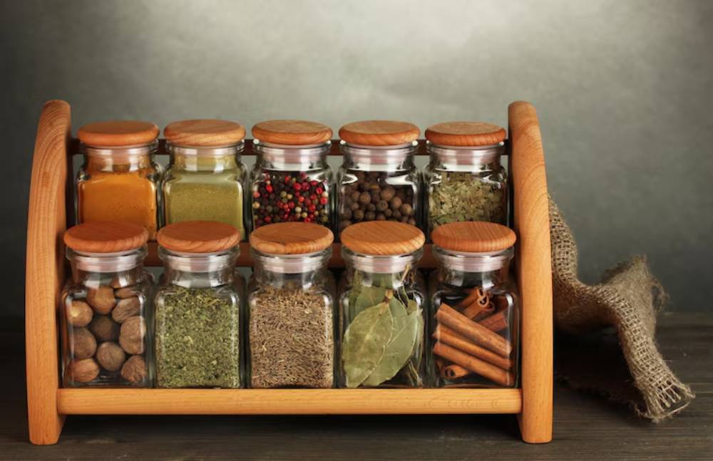A wooden spice rack holding twelve glass jars filled with various spices, set against a muted gray background.