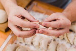 A pair of hands expertly pleating a stuffed pasta, surrounded by uncooked pasta on a wooden surface.