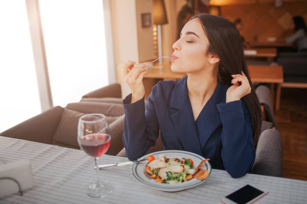 A person in a navy blue blazer enjoys a meal of chicken and vegetables, accompanied by a glass of red wine at a cozy restaurant.