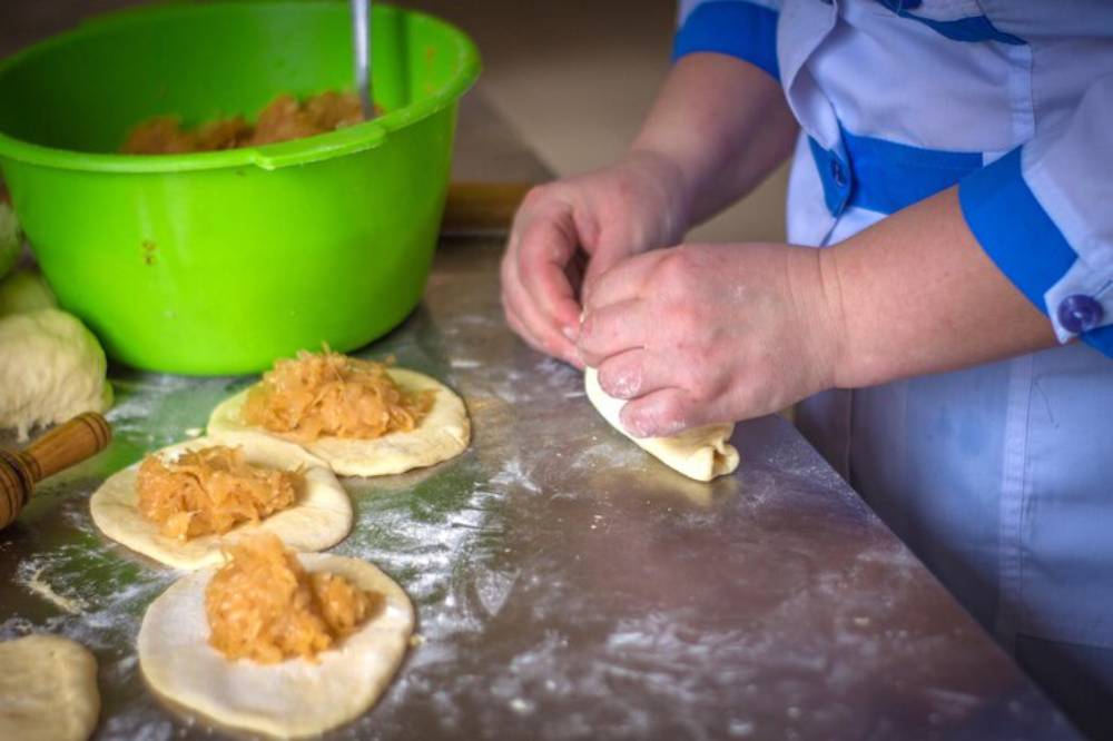 A cook prepares dough filled with filling, folding it carefully, with a green bowl of filling in the background and flour on the surface.