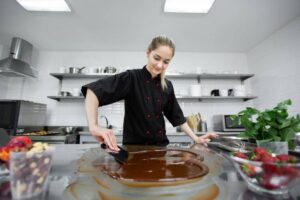 A chef in a black coat smooths melted chocolate on a shiny surface in a modern kitchen, surrounded by fresh strawberries and nuts.
