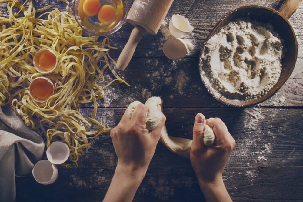 Aerial view of hands kneading dough, surrounded by pasta, eggs, flour, and a rolling pin on a rustic wooden table.