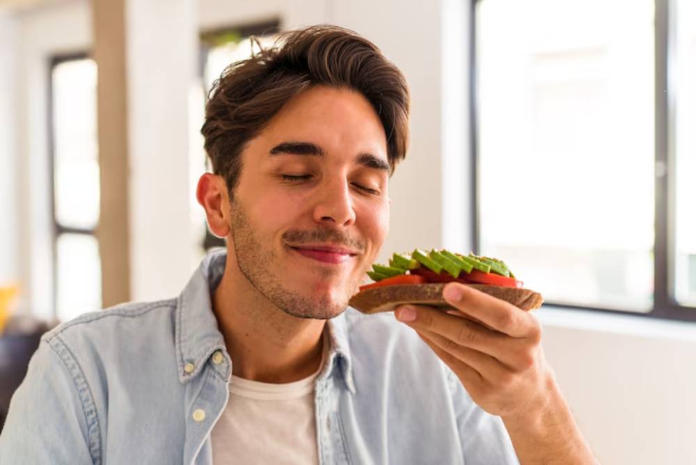 A person holding a colorful avocado toast topped with green avocado slices and red tomatoes, sitting in a bright, airy café.