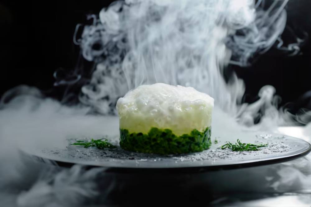 A gourmet dish featuring a layered green base topped with a creamy white foam, surrounded by swirling smoke on a sleek black plate.