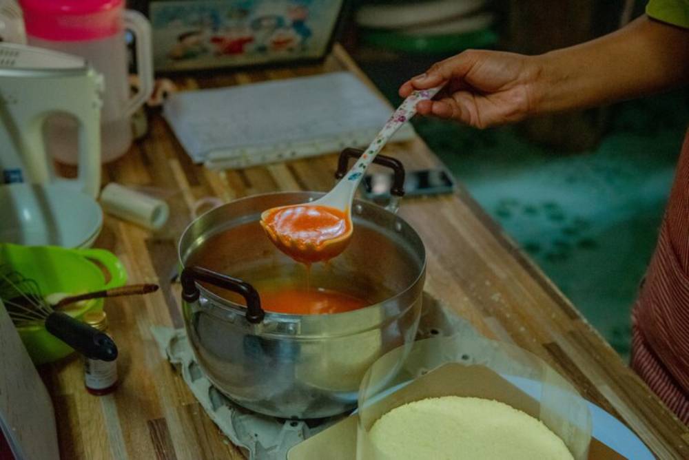 A hand holds a spoonful of vibrant orange sauce above a pot on a wooden countertop, with baking tools scattered nearby.