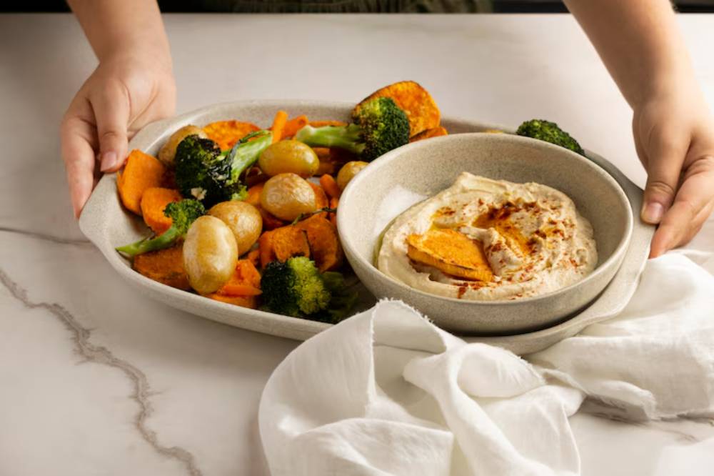 A person holds a platter of roasted vegetables and a bowl of hummus, showcasing a colorful and healthy meal.