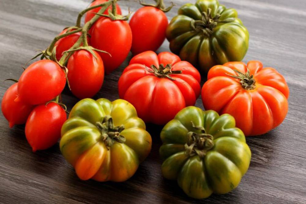 A variety of tomatoes: red grape tomatoes alongside several green and ripe heirloom tomatoes on a wooden surface.