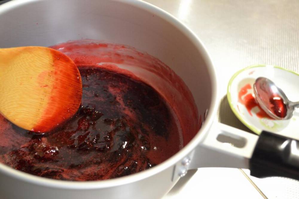 A wooden spoon stirs a thick, dark red berry sauce in a pot, with a small dish and spoon beside it, stained with sauce remnants.