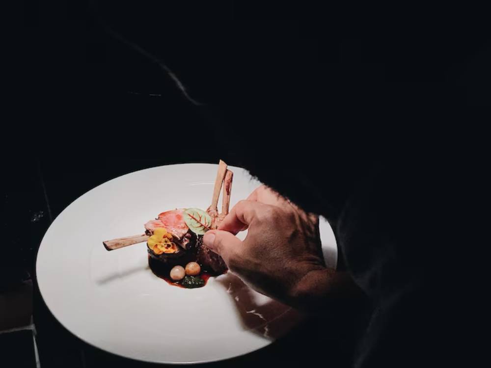 A chef delicately garnishes a gourmet dish featuring meat, vegetables, and sauces on a white plate in low light.
