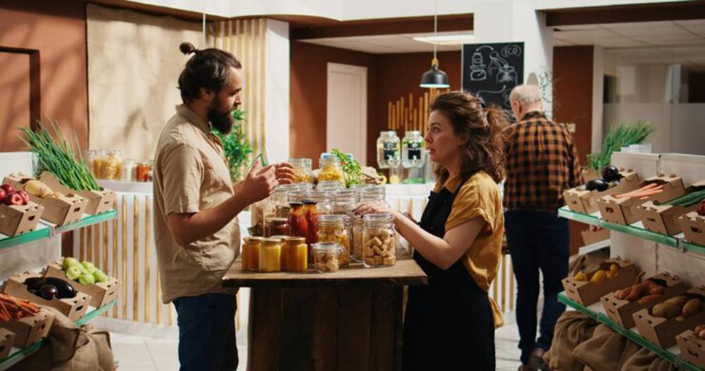A man and woman standing examines jars of food on a wooden table in a cozy market filled with fresh produce and home-made goods.