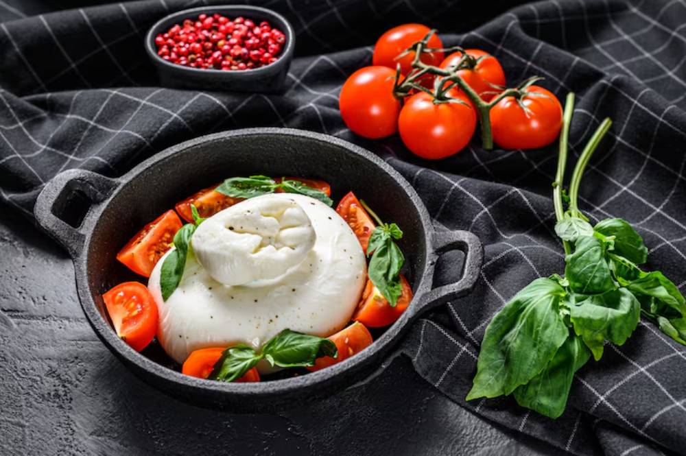 A cast iron dish filled with fresh tomatoes, basil, and a creamy burrata cheese, surrounded by vibrant cherry tomatoes on a checked cloth.
