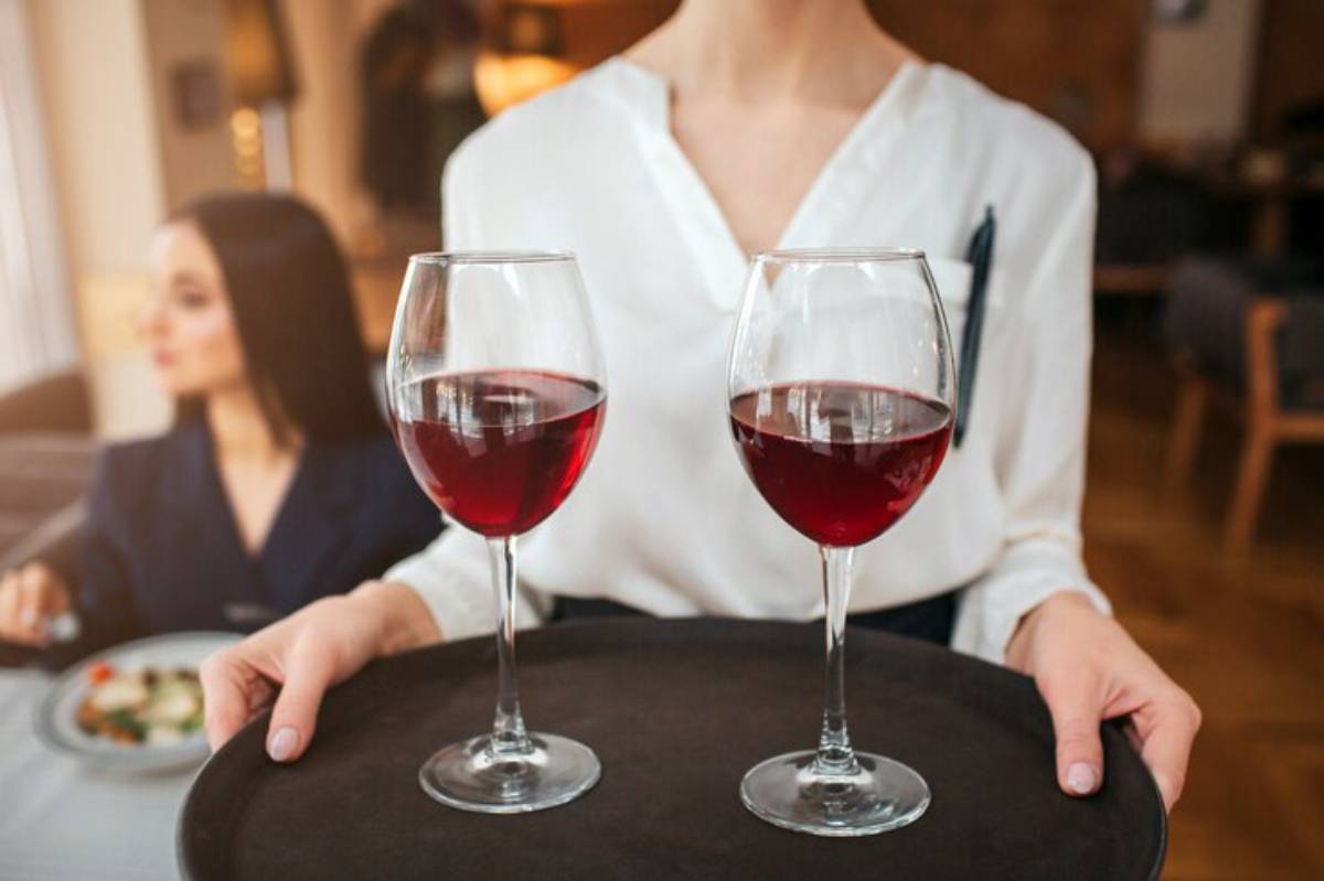 A server in a white blouse presents a tray with two glasses of red wine in an elegantly styled restaurant setting.