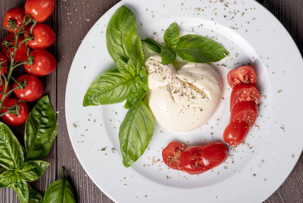 Fresh burrata cheese garnished with basil leaves, paired with sliced tomatoes, and sprinkled with herbs on a white plate.