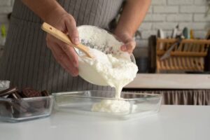 A person in an apron pours fluffy cream from a glass bowl into a clear dish, with cookies in the foreground on a white countertop.
