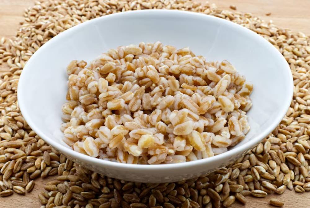 A white bowl filled with Farro , surrounded by raw Farro kernels on a wooden surface.