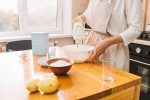 A person in an apron uses a hand blender to preparing foam in a white bowl on a wooden table, with sugar and lemons nearby.