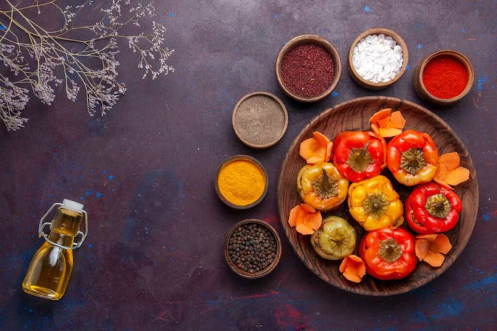 A wooden plate holds colorful stuffed bell peppers, surrounded by bowls of spices, salt, and a glass bottle of oil on a dark background.
