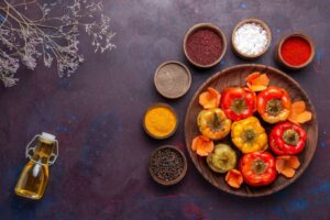 A wooden plate holds colorful stuffed bell peppers, surrounded by bowls of spices, salt, and a glass bottle of oil on a dark background.