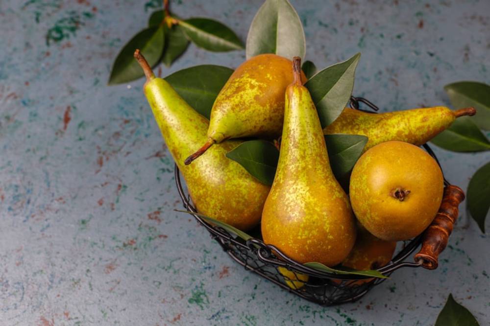 A wire basket filled with ripe yellow-green pears surrounded by green leaves, resting on a textured blue background.
