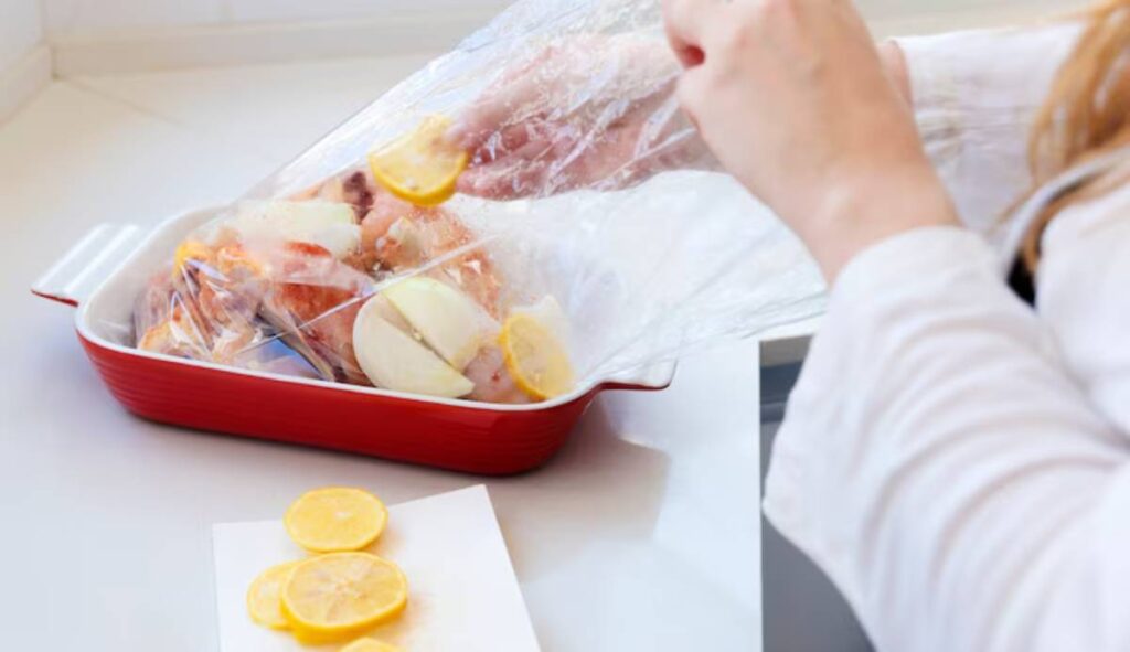 A person adds lemon slices to seafood in a red baking dish, covered with plastic wrap, on a kitchen countertop.